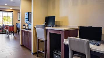 A hotel room with a desk, computer, and coffee machine. There are chairs and a window with a view of trees.