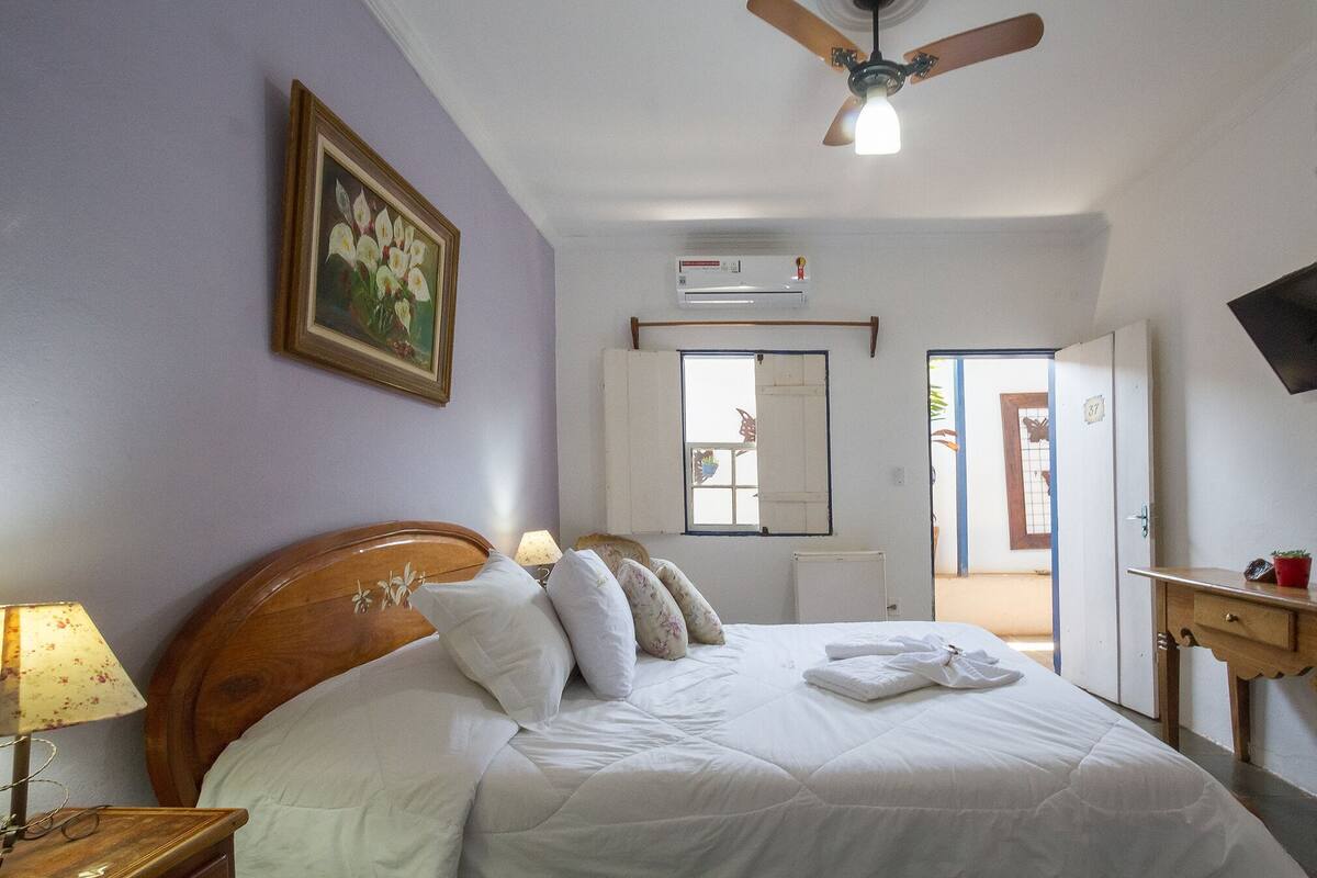 A bedroom with a bed, a ceiling fan, an air conditioning unit, a wooden nightstand, a lamp, and a painting on the wall.