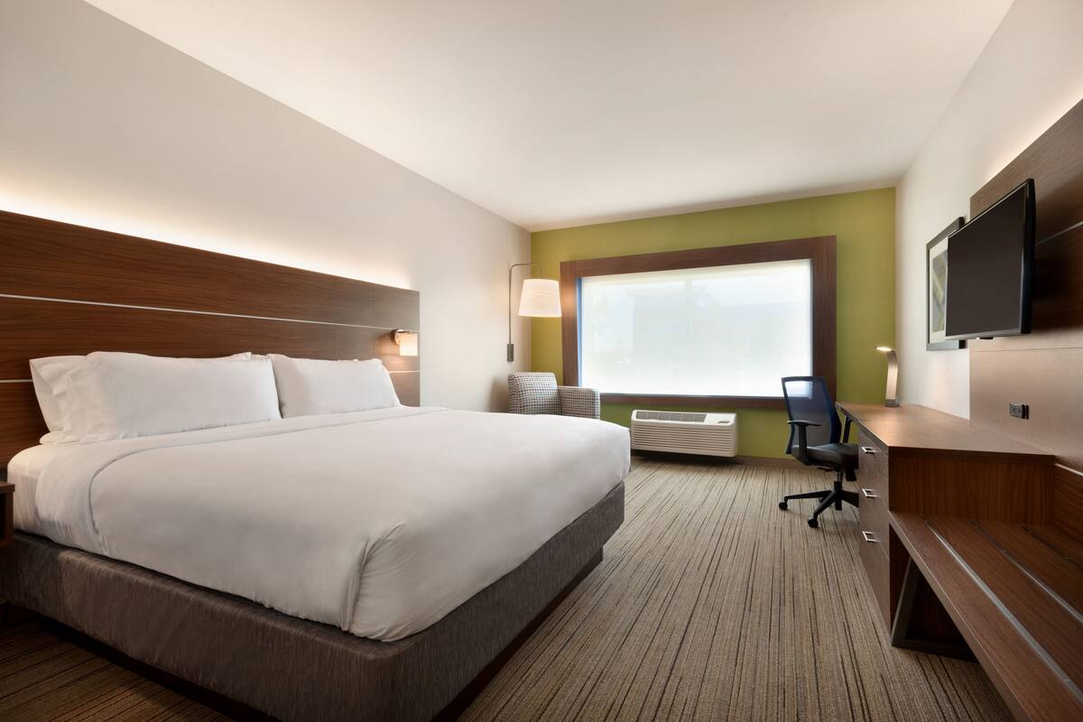 Ein Hotelzimmer mit einem großen Bett, einem Schreibtisch mit Stuhl, einem Flachbildfernseher, einem Fenster mit Jalousien und einer Wandlampe.