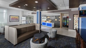 A lobby with a brown sofa, grey armchairs, a small round table, and a vending machine. - Holiday Inn Express & Suites West Des Moines - Jordan Creek by IHG (West Des Moines)