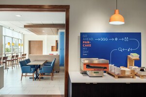 Un comedor moderno con un televisor de pantalla plana mostrando un menú de desayuno, una cafetera y un conjunto de mesas y sillas.