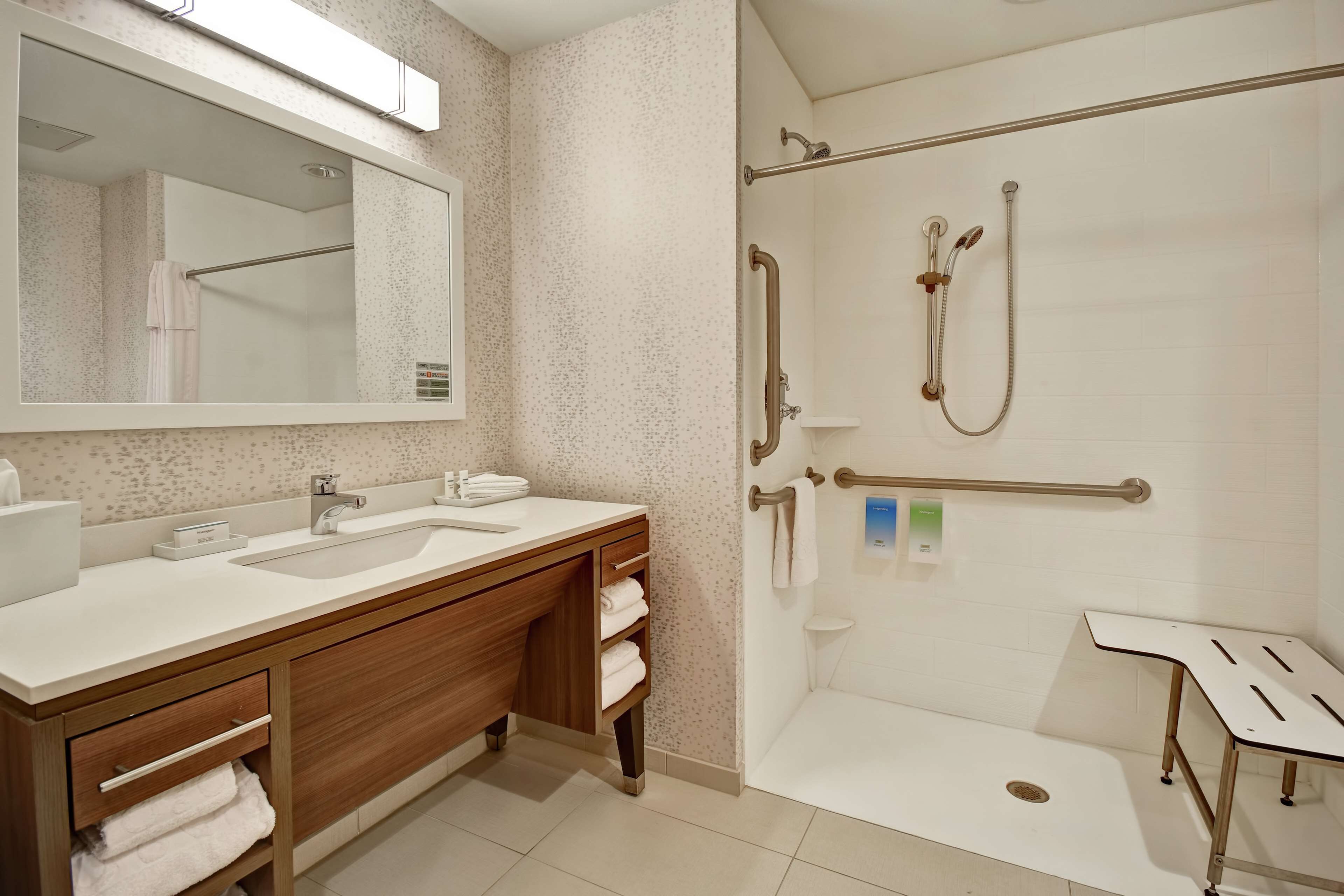 A modern bathroom with a large mirror, two sinks, wooden vanity, and a walk-in shower with grab bars.