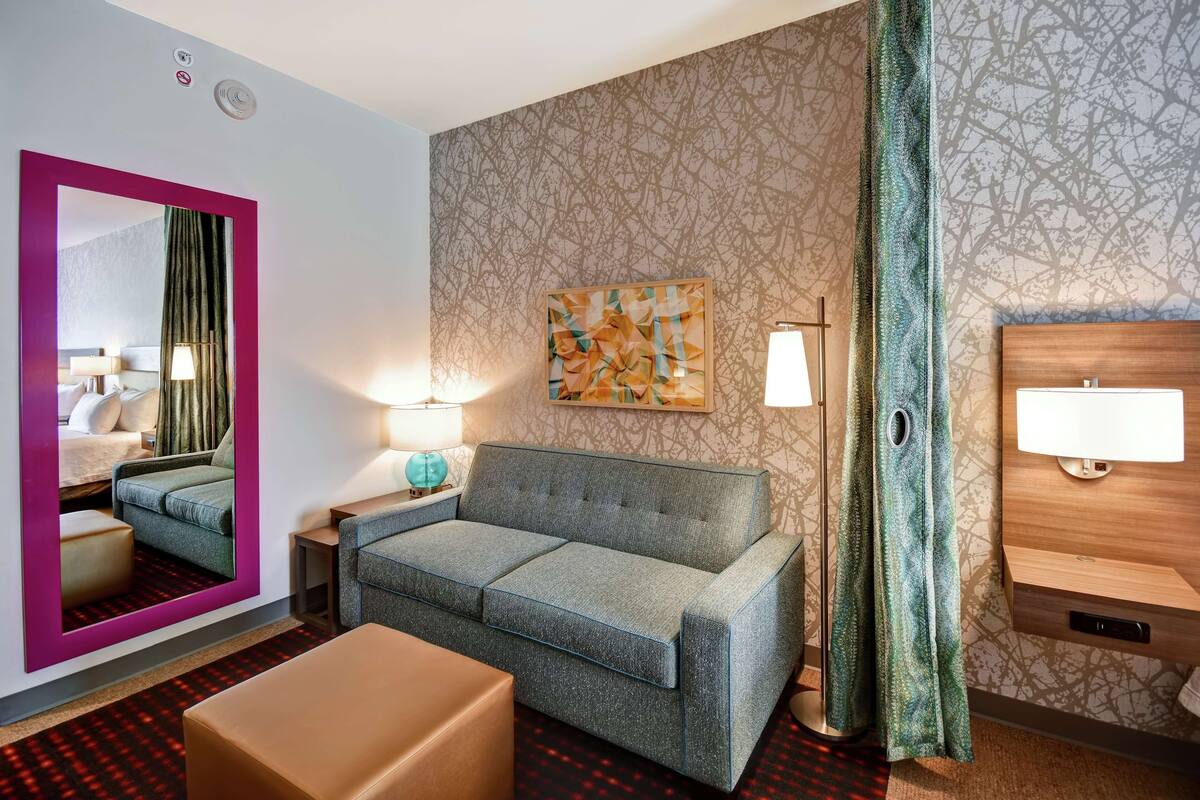 A hotel room with a grey sofa, a small table, a mirror with a pink frame, a wall-mounted lamp, and a patterned wallpaper.