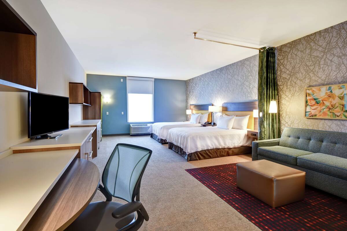 A hotel room with a desk, two beds, a sofa, a TV, and a large window.