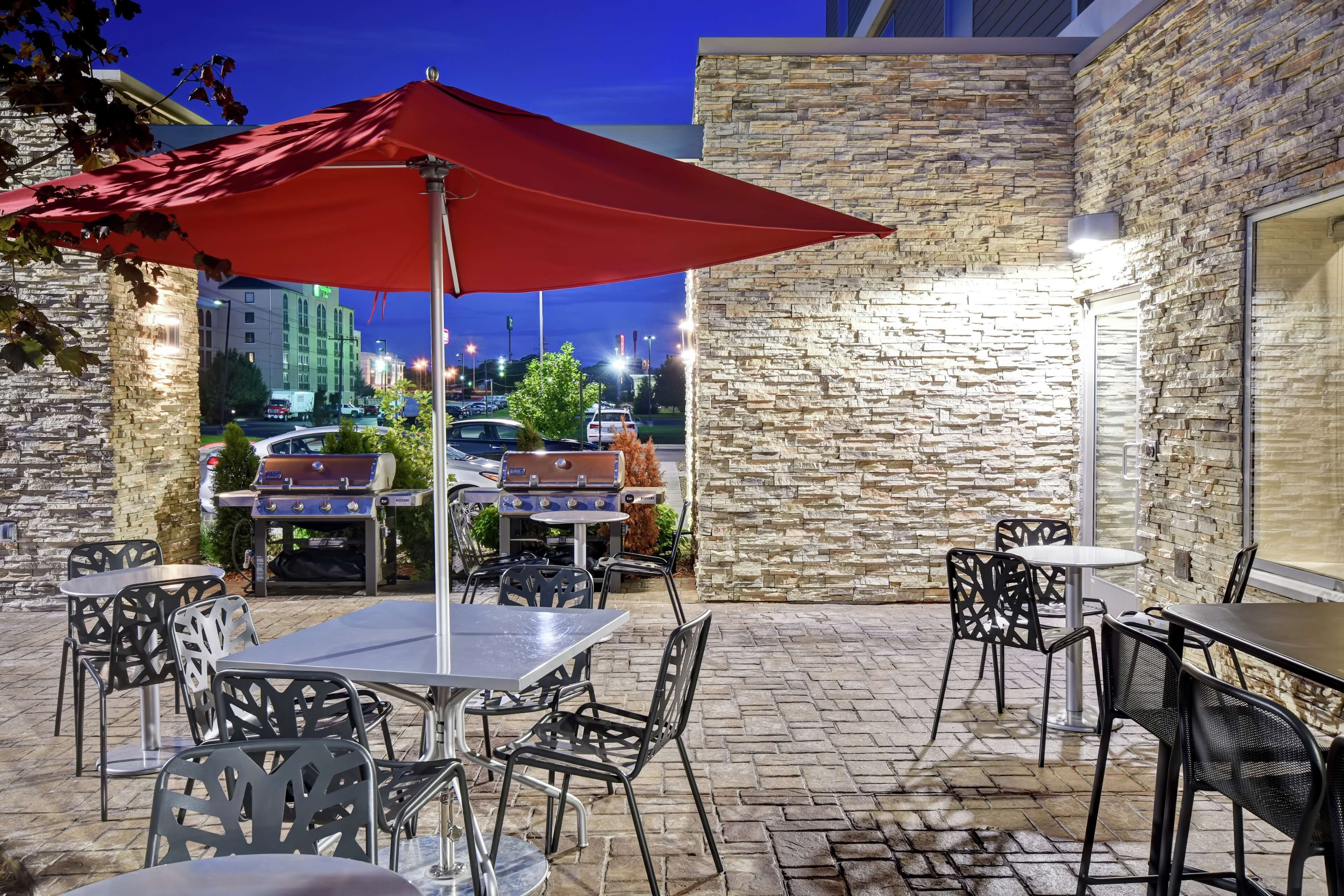 Un patio exterior con paredes de piedra, mesas y sillas, que incluye una sombrilla roja y parrillas para barbacoa.