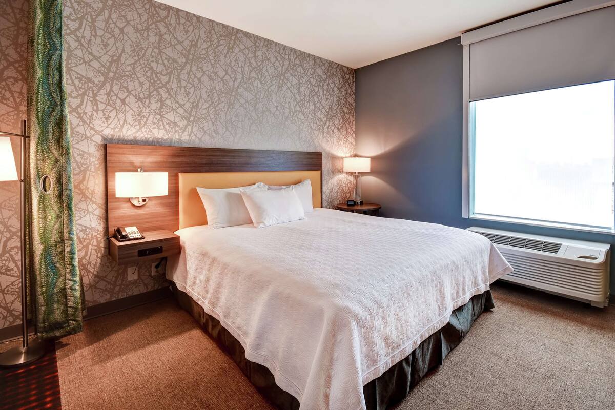 A hotel room with a large bed, two bedside lamps, a nightstand with a phone, a window with a view, and a wall with a patterned wallpaper.