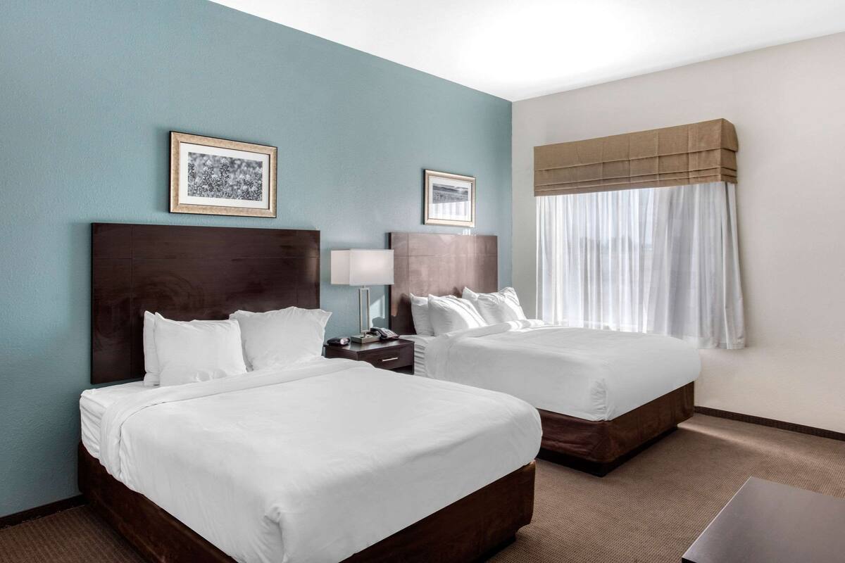 A hotel room with two beds, a nightstand, a lamp, and a window with curtains.