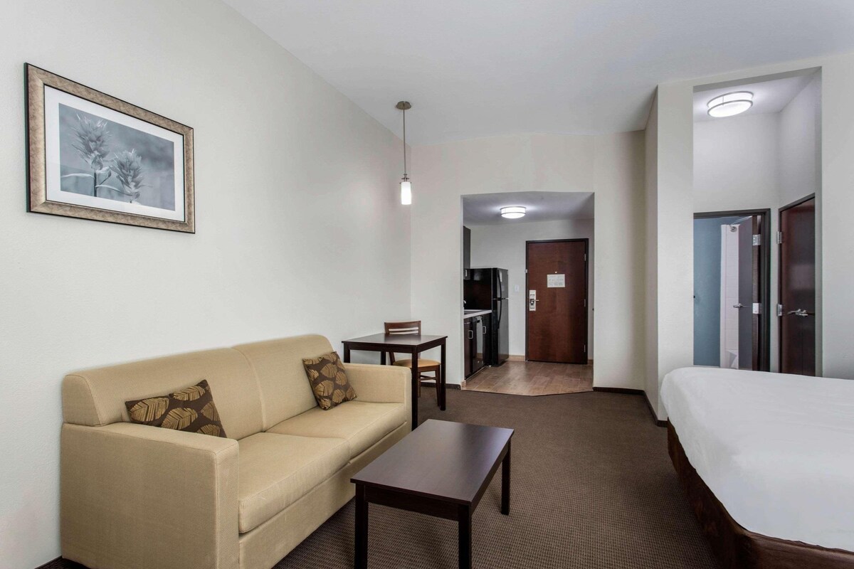 A hotel room with a sofa, a small table, a bed, a kitchenette, and a bathroom visible through an open door.