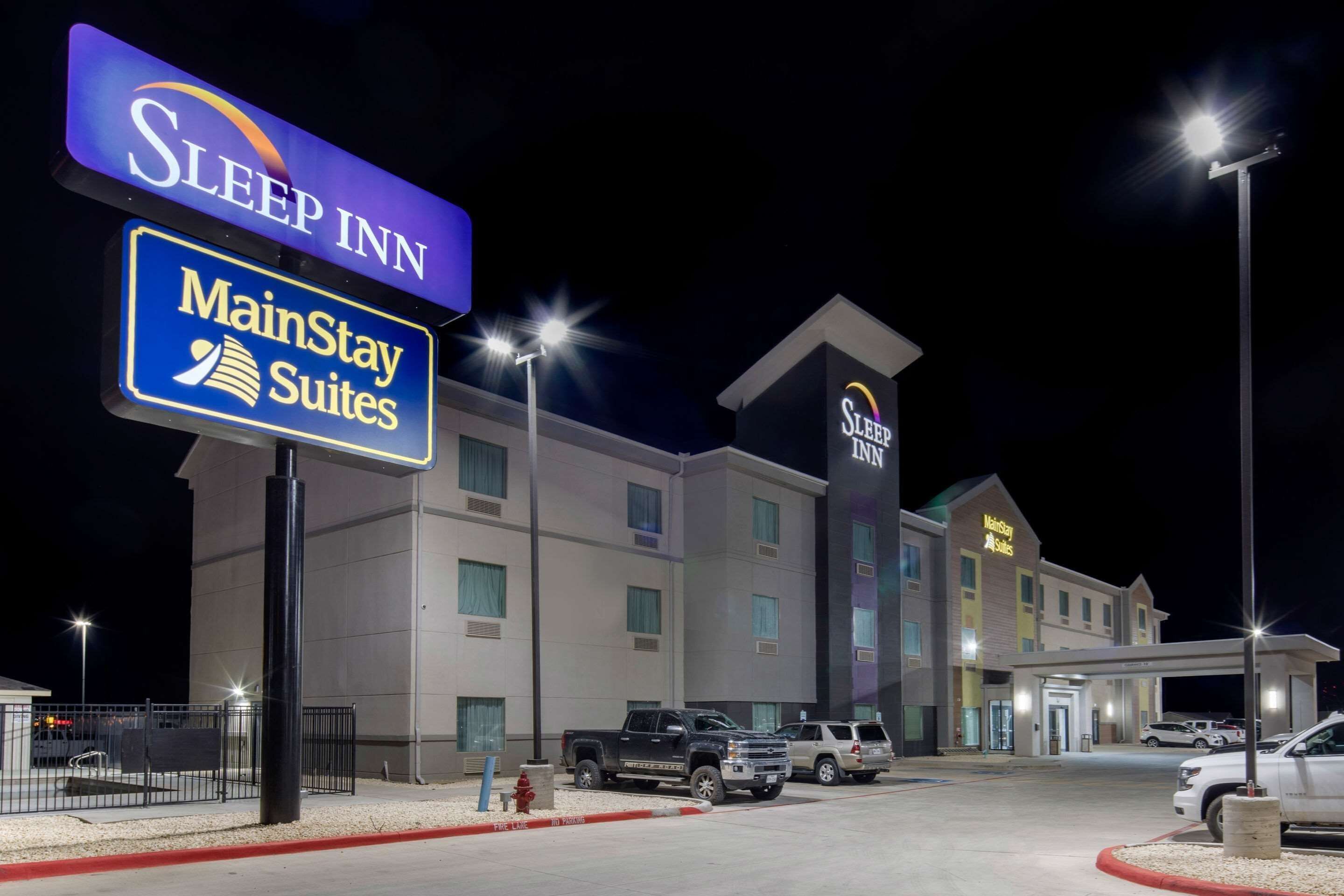 Un bâtiment de la chaîne Sleep Inn MainStay Suites la nuit, avec une enseigne lumineuse, un parking et une entrée fermée par un portail.