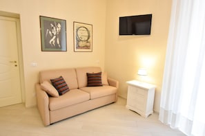 Ett vardagsrum med en beige soffa, två kuddar, ett litet vitt sidobord med en lampa och en platt-TV som är monterad på väggen.
