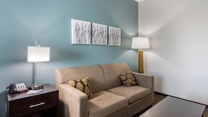 A hotel room with a beige sofa, two pillows, a small table, a nightstand with a lamp, a phone, and a wall-mounted artwork. - Sleep Inn Big Spring North I-20 (Big Spring)