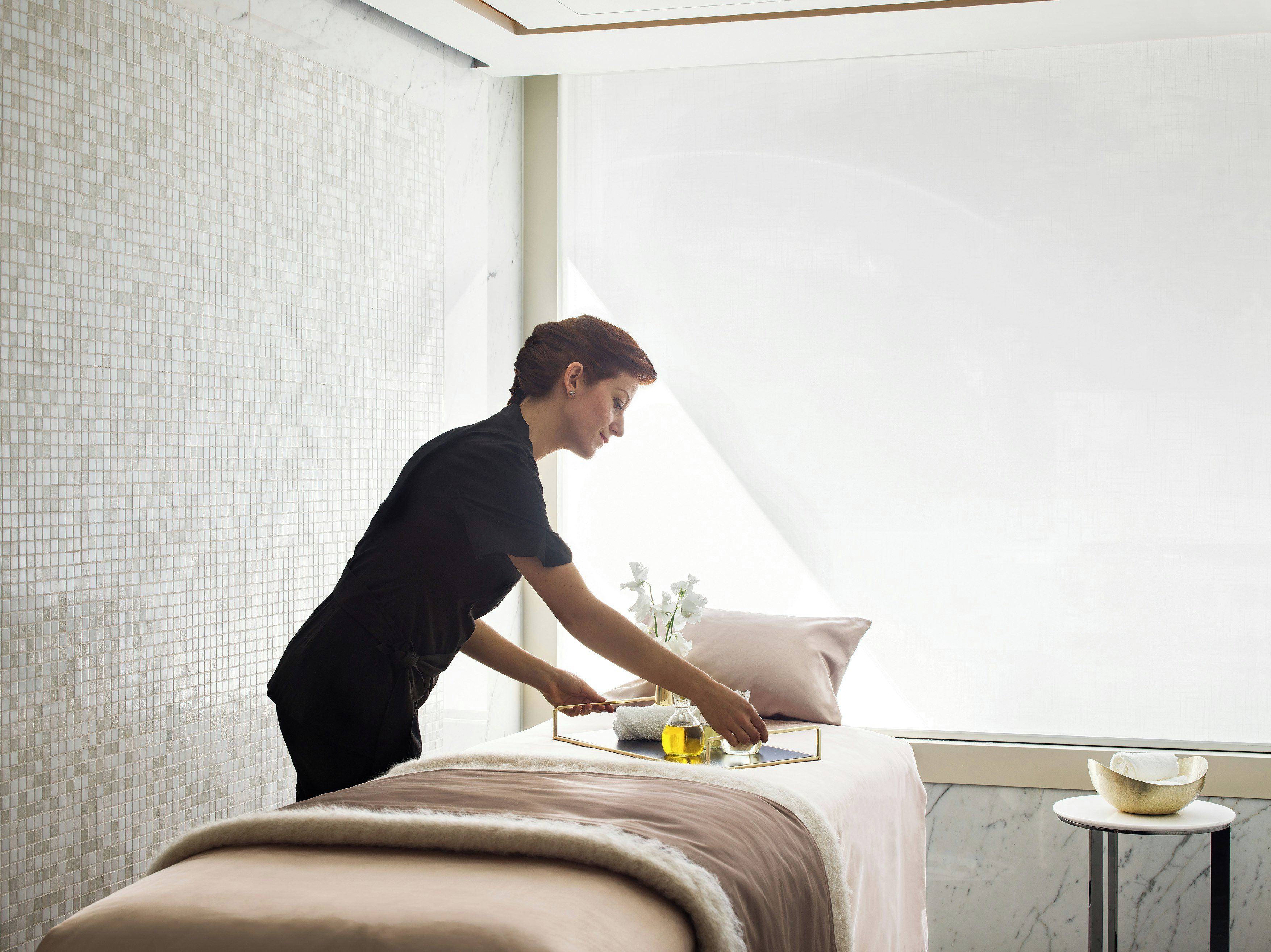 A person attending to a massage table with a towel and a glass bottle, in a room with a tiled wall and a window.