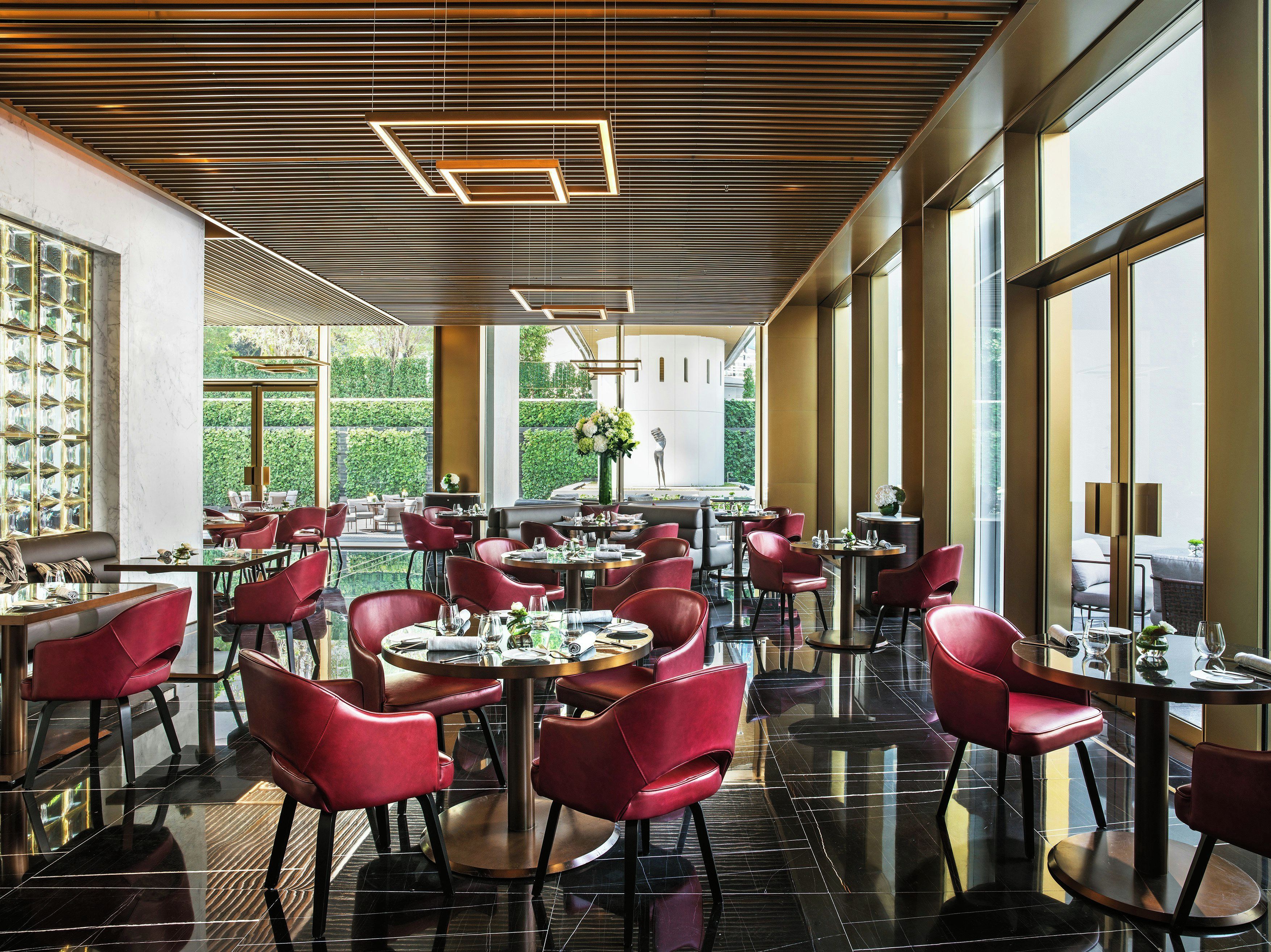 Een modern restaurant met ronde tafels en rode stoelen, grote ramen en een plafond met lineaire verlichting.