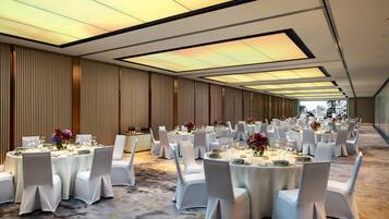 A banquet hall with round tables, white chairs, and a city view through large windows.