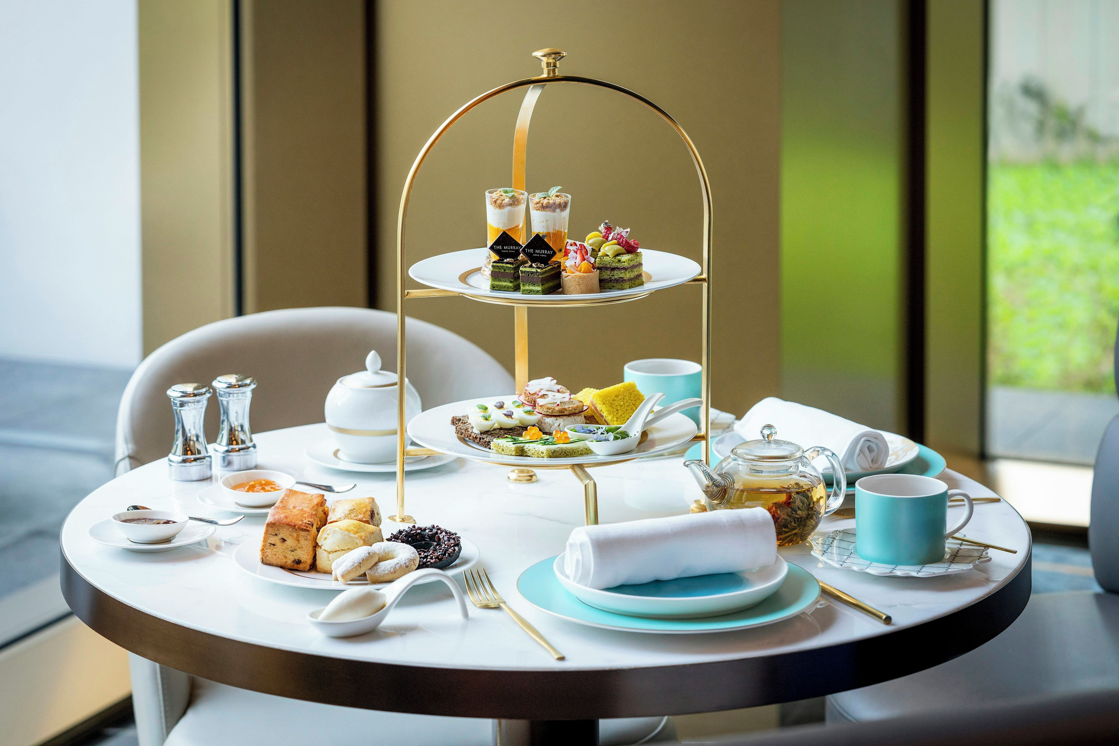 Une table ronde sur laquelle repose un présentoir à étages contenant divers gâteaux et desserts, ainsi que des tasses à thé et à café, et une théière.