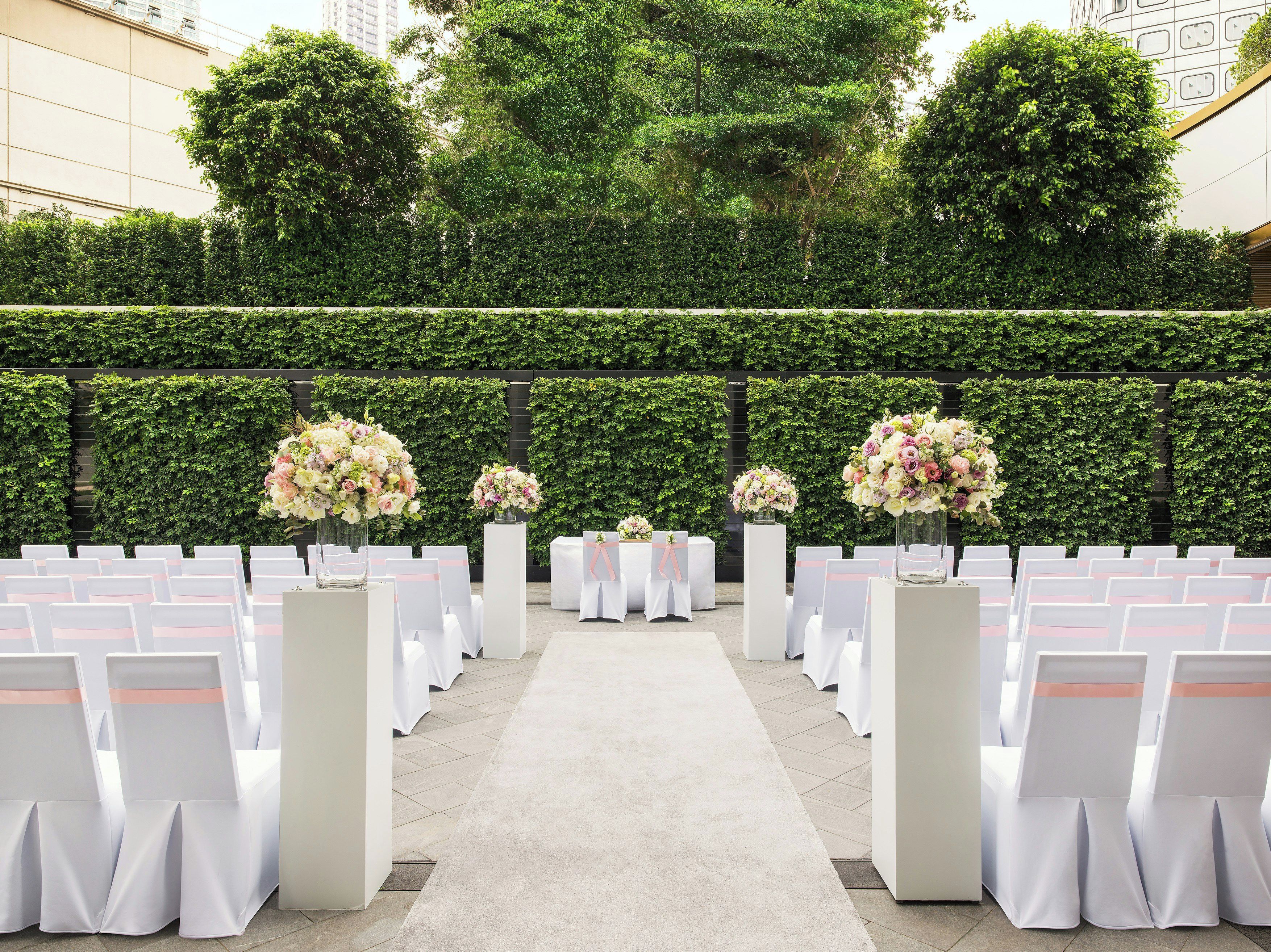 A formal outdoor event setup with rows of white chairs and tables, floral centerpieces, and a hedge wall in the background.
