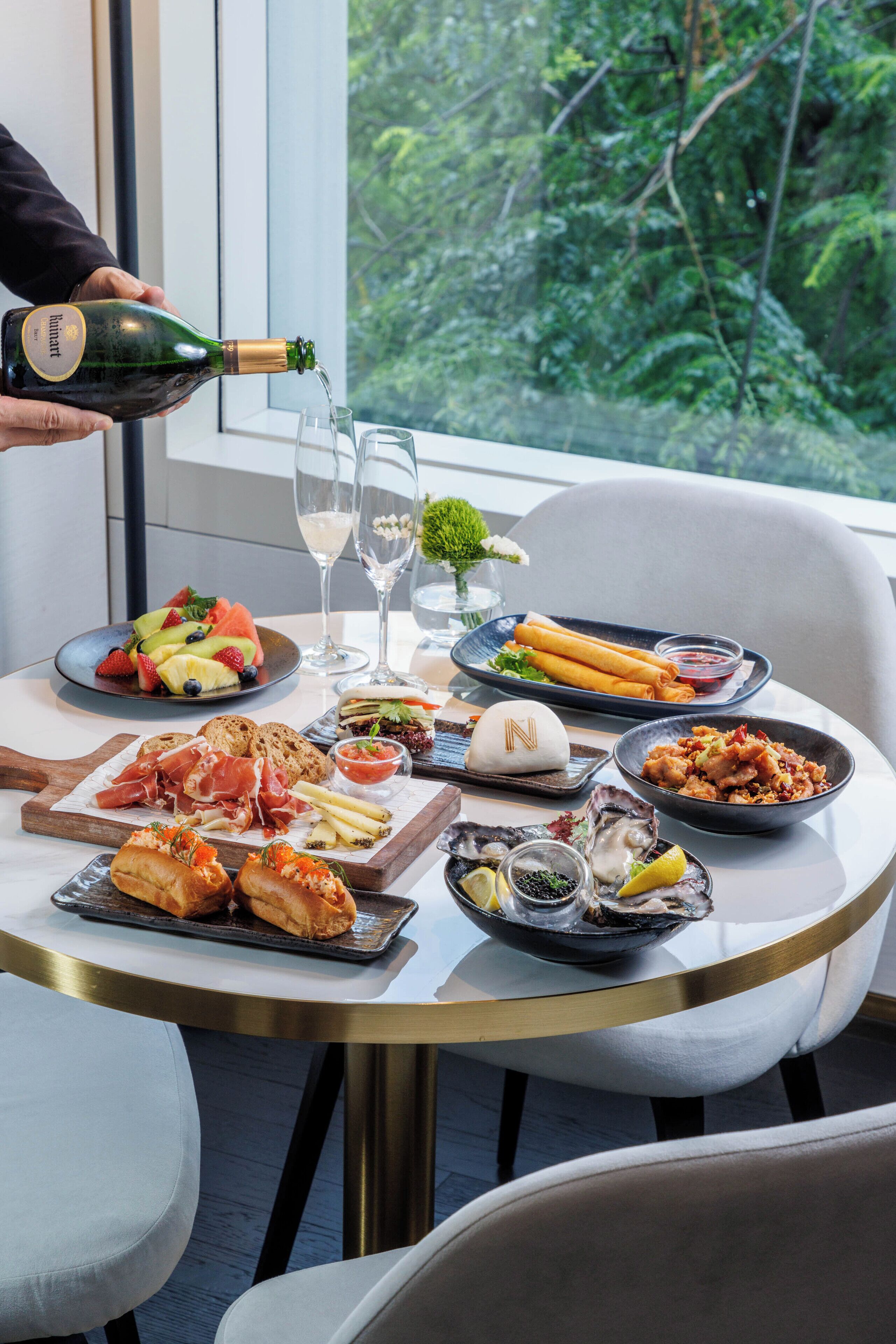A table set with various food items, a bottle of champagne being poured, and a scenic view through a window.