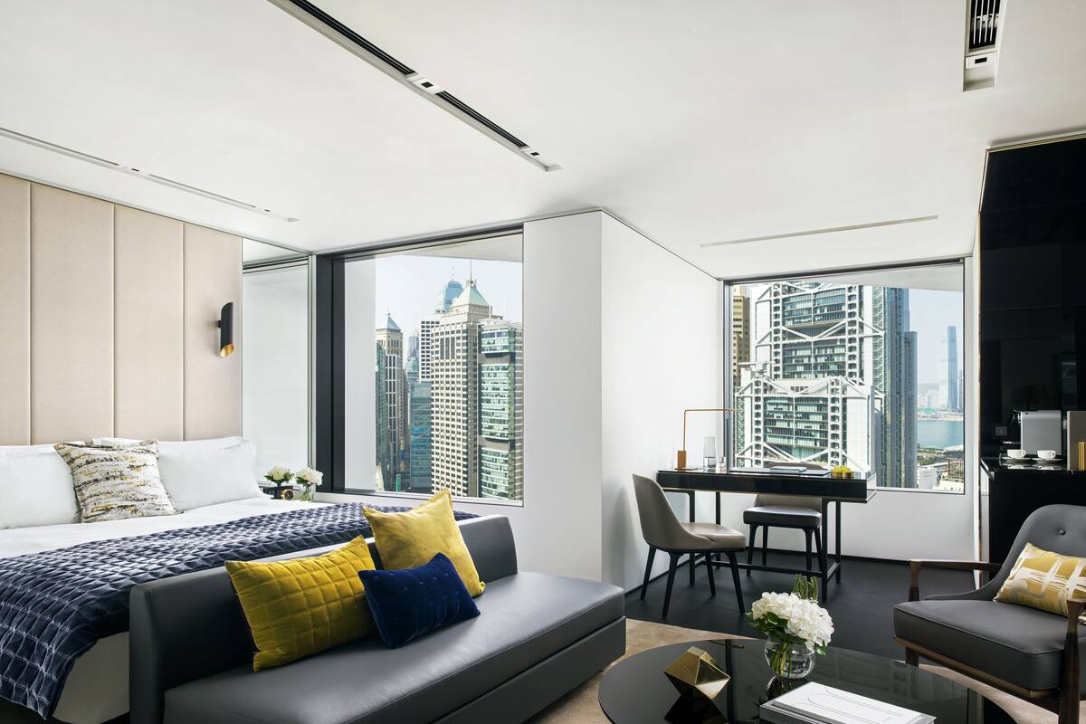 A modern hotel room with a large bed, a sofa, a dining area, and a view of city skyscrapers.