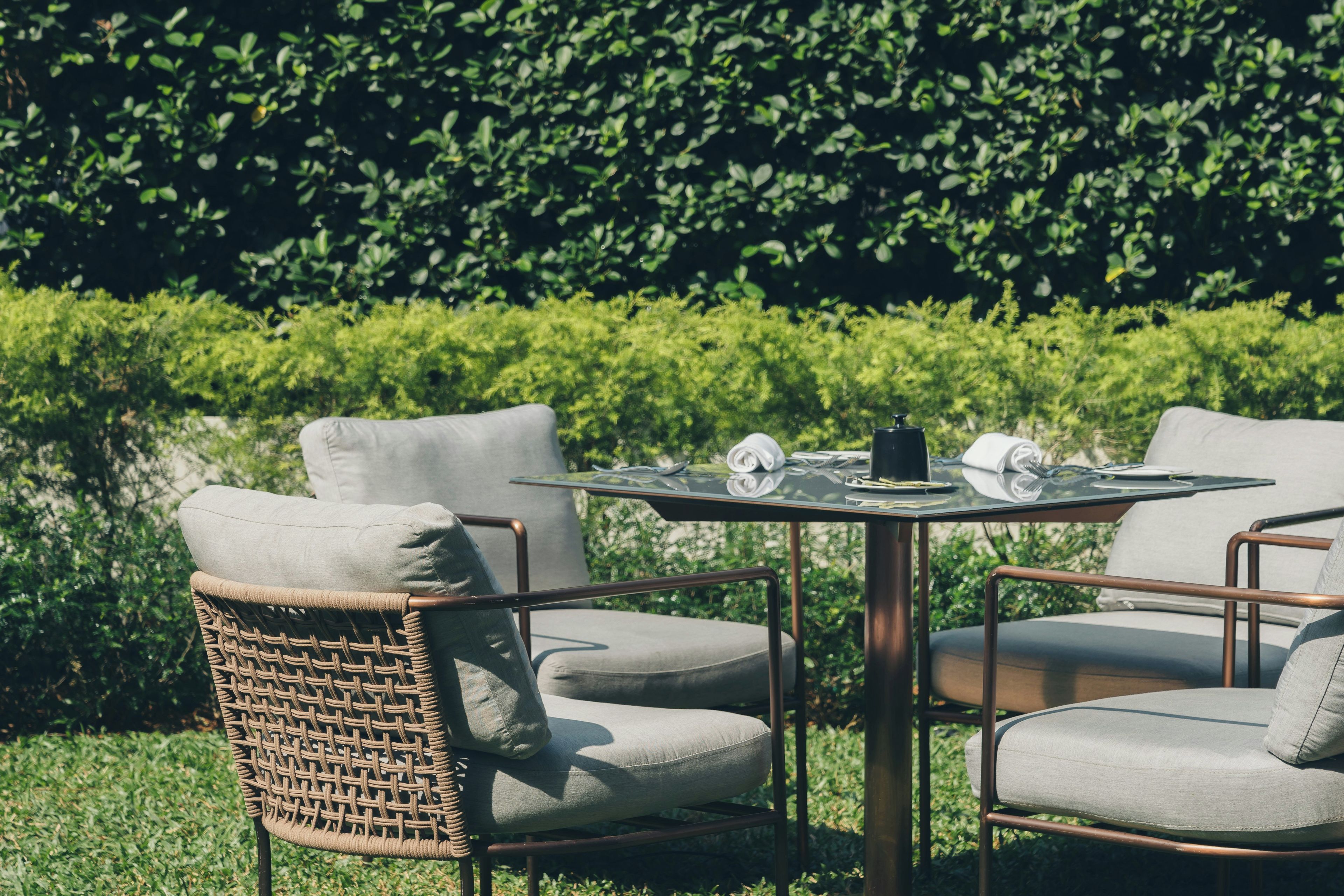 A round table with two chairs, a small black object on the table, and a hedge in the background.