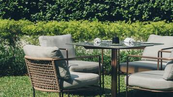 A round table with two chairs, a small black object on the table, and a hedge in the background.