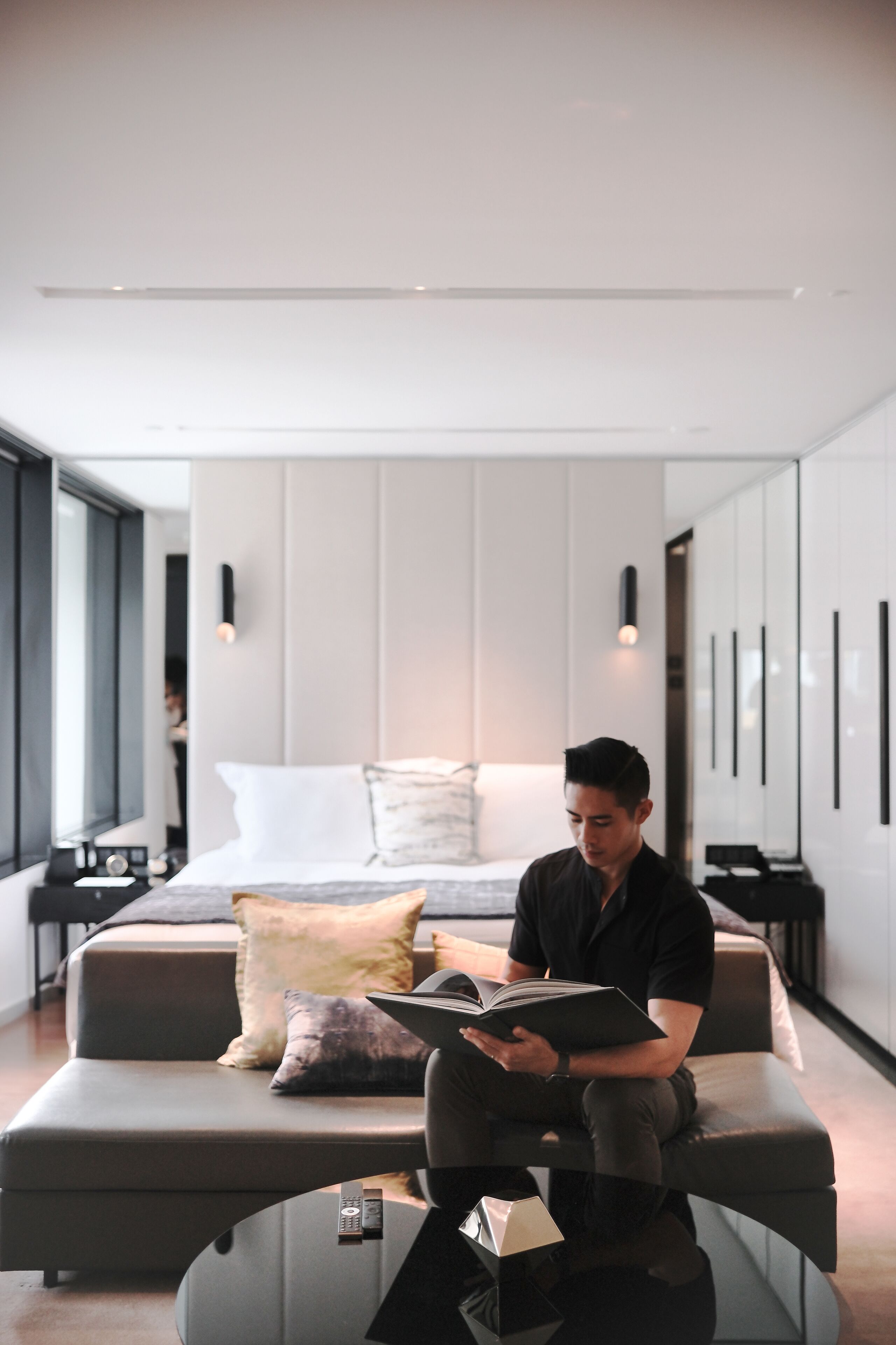 A modern hotel room with a sofa, a glass coffee table, and a bed with pillows.