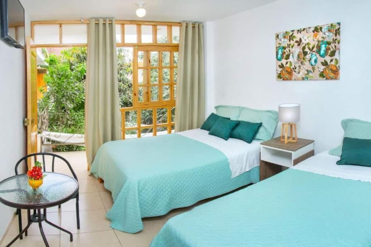 A room with two beds, a small table with a chair, a window with curtains, and a painting on the wall.