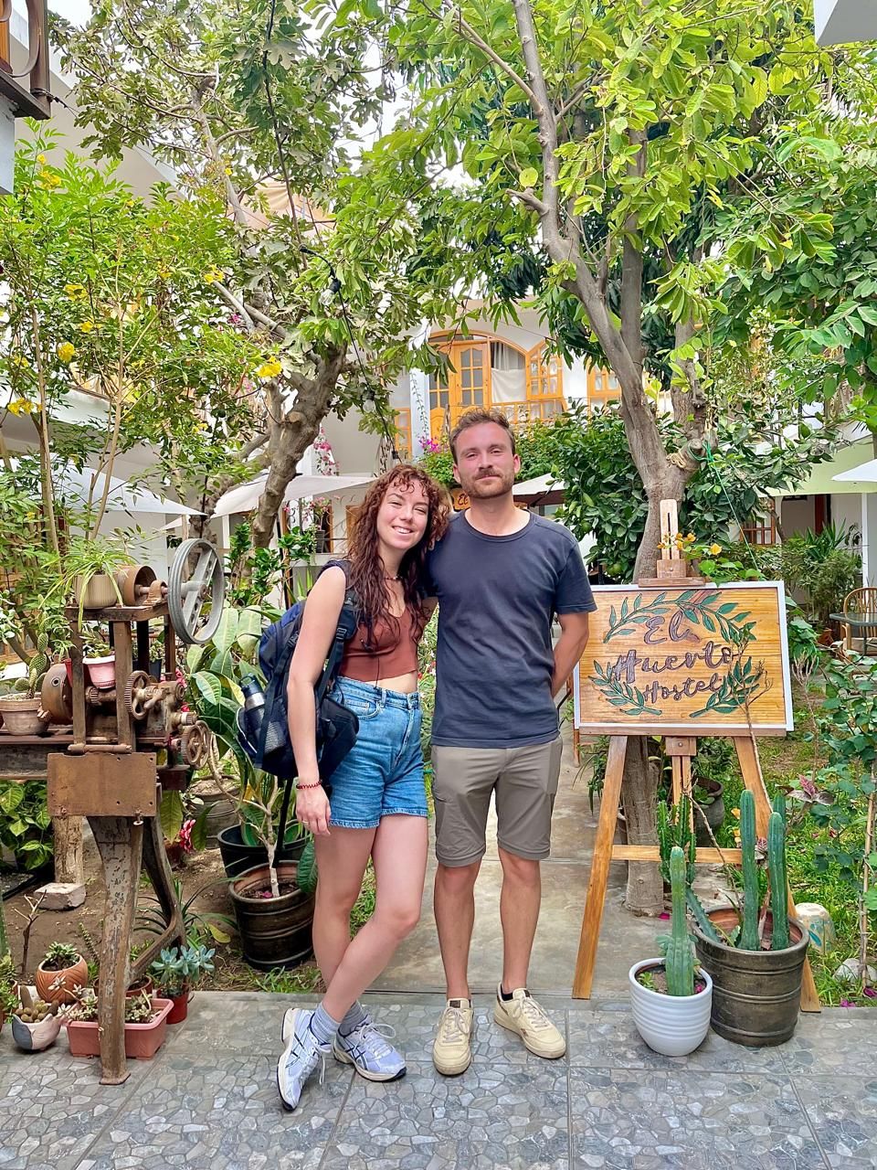 Una pareja de pie en un patio interior con un letrero que dice 'El Huerto Hostel'.