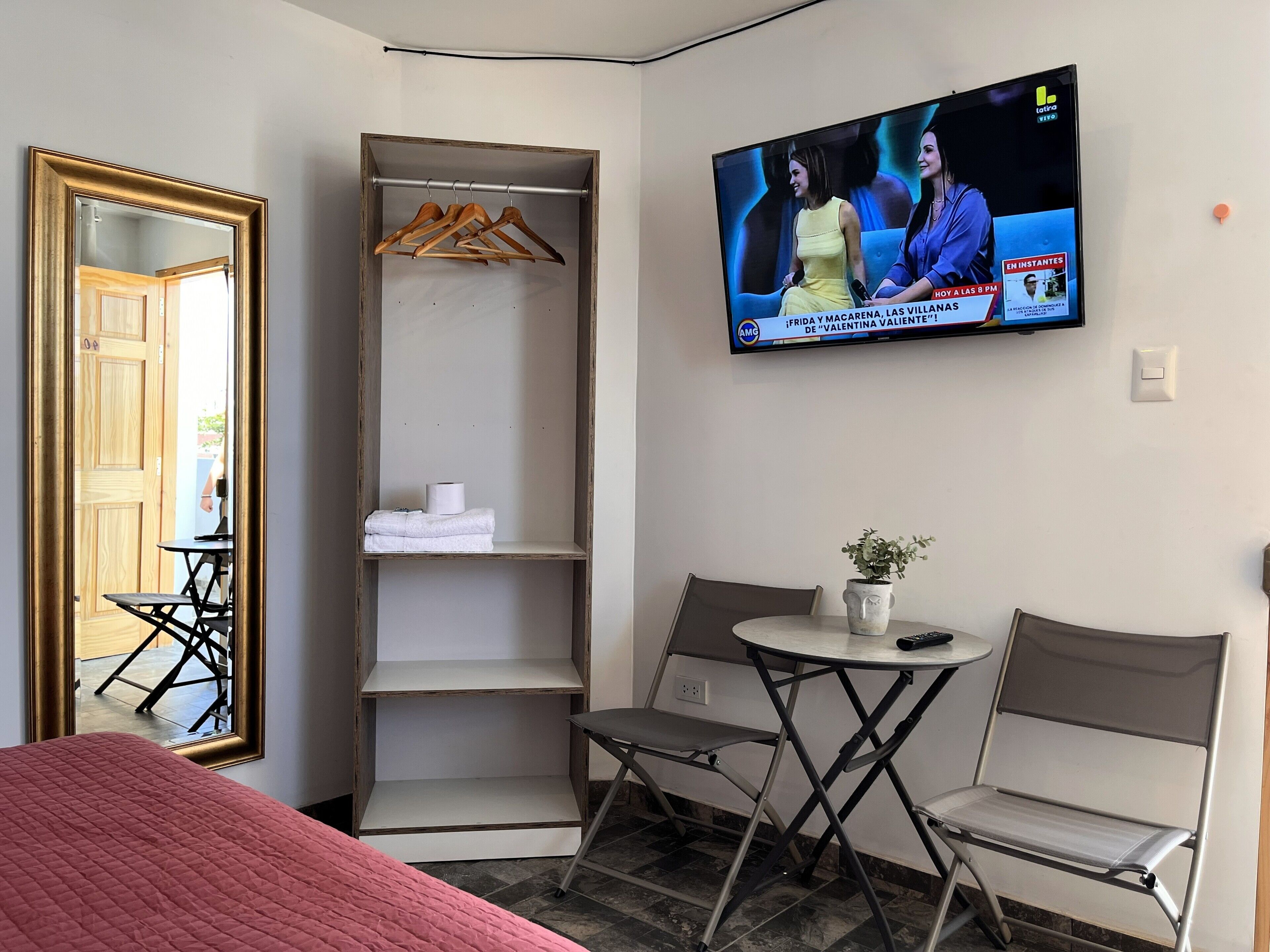 Habitación de hotel con cama, televisión montada en la pared, armario y una pequeña mesa con silla.