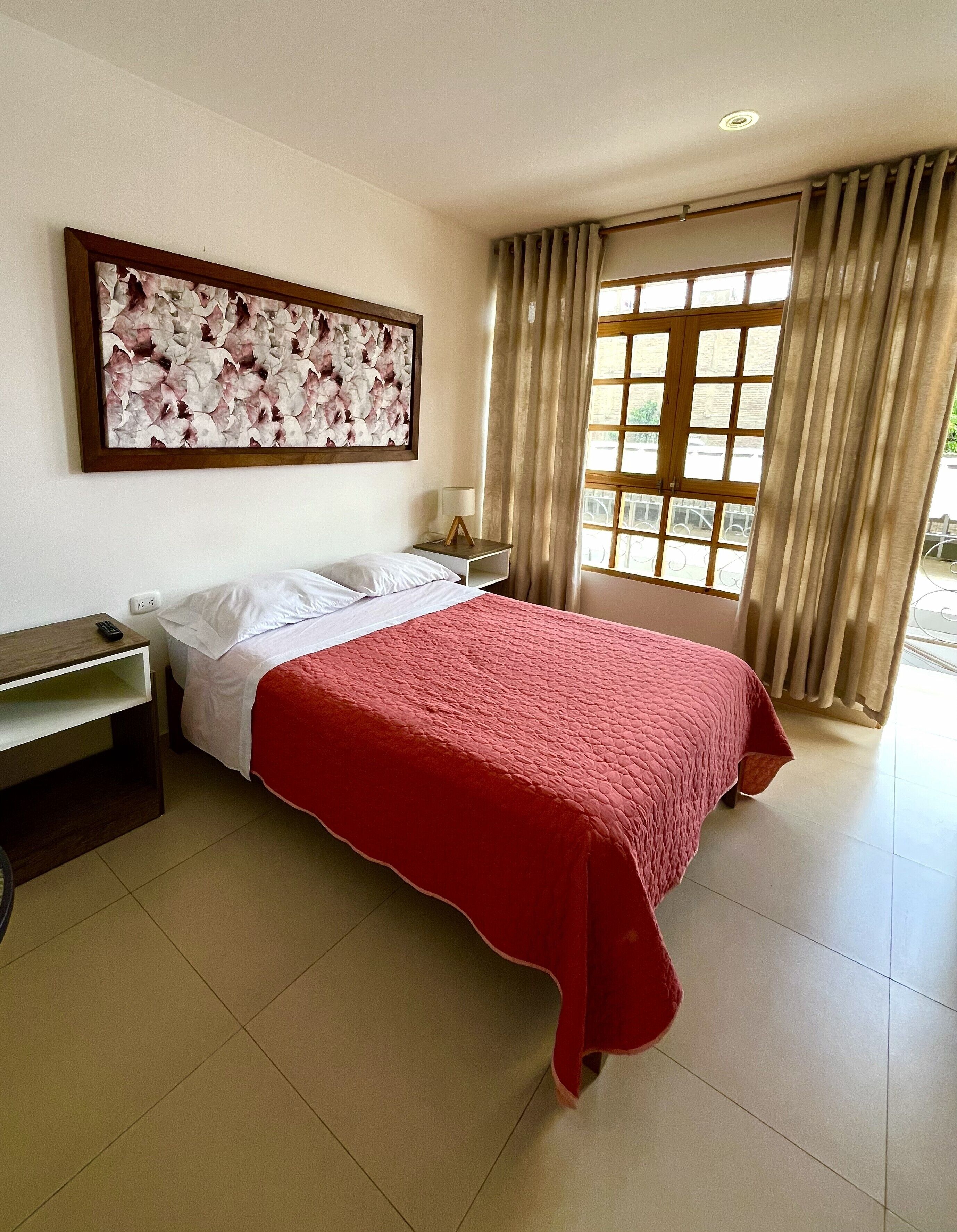 Una habitación de hotel con cama, mesita de noche, lámpara, un cuadro en la pared y ventana con cortinas.