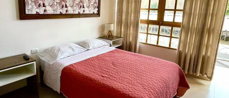 Una habitación de hotel con cama, mesita de noche, lámpara, un cuadro en la pared y ventana con cortinas.