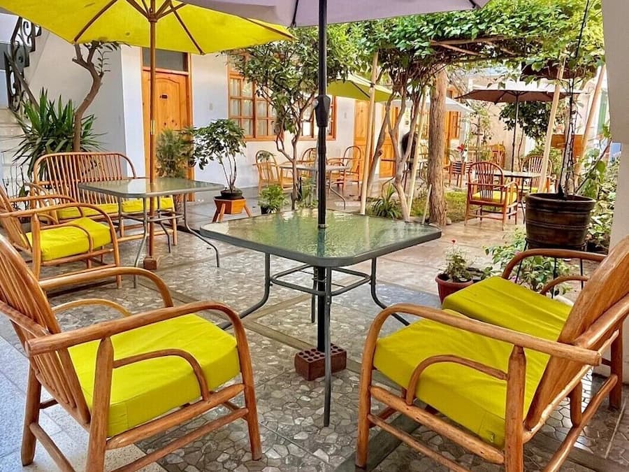 Un patio al aire libre con una mesa de vidrio rodeada de sillas de mimbre con cojines amarillos, bajo un paraguas grande.