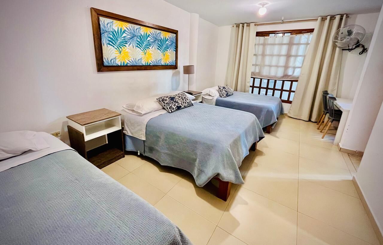 A hotel room with two beds, a small desk, a chair, a fan, and a window with curtains.