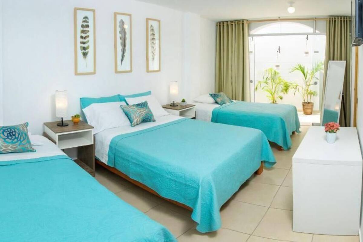 A hotel room with three beds, each with a teal bedspread and matching pillows. There are bedside tables with lamps, a large mirror, and a view of the outdoors through a glass door.