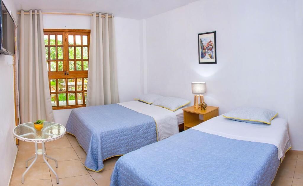 Quarto com duas camas, uma mesa pequena com uma planta, uma janela com cortinas, uma mesinha de cabeceira com abajur e um quadro na parede.