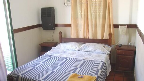 A single bed with a striped bedspread and pillows, a bedside table, a nightstand, a wall-mounted heater, and a window with curtains. - Prisma Apart Hotel (La Rioja)