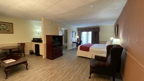 Une chambre d’hôtel avec un lit, un bureau, une télévision, une chaise et une salle de bain visible à travers une porte ouverte.