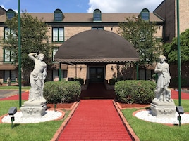 Un edificio con un sendero de ladrillo rojo que conduce a una entrada cubierta, flanqueado por estatuas y follaje.