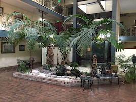Um amplo átrio interno com um jardim central, apresentando plantas em vasos grandes, uma estátua e uma fonte.