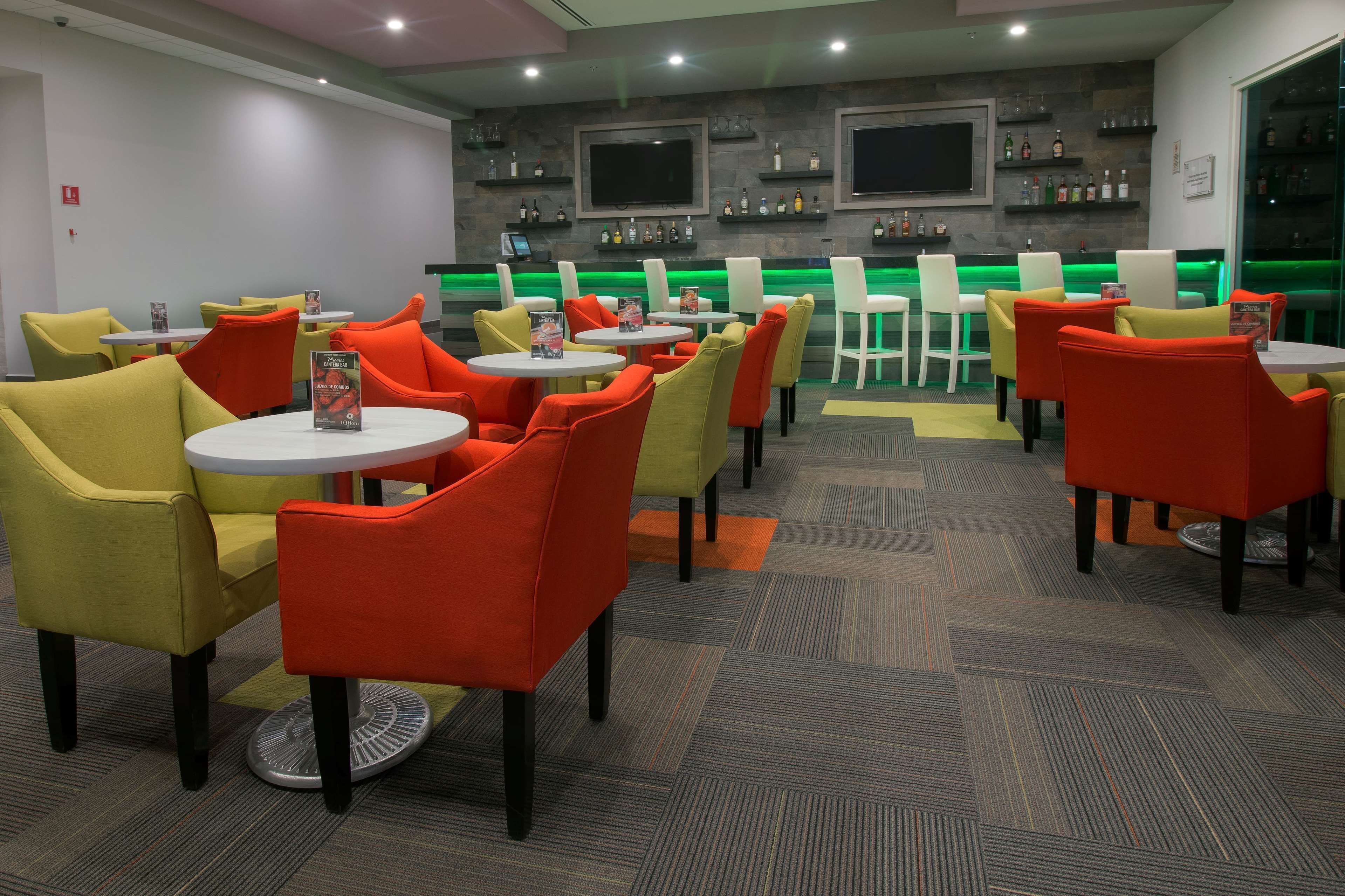 Un restaurant moderne avec des tables rondes et des chaises aux couleurs vives, un bar avec un éclairage vert et un téléviseur fixé au mur.