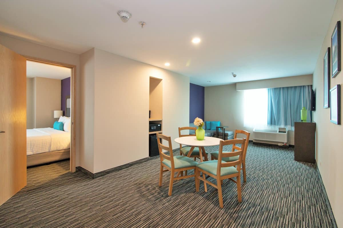 Une chambre d’hôtel comprenant un lit, une table à manger avec des chaises, un petit coin cuisine et une grande fenêtre avec des rideaux.