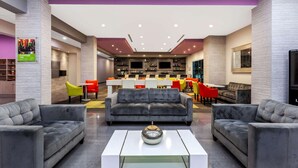 Un hall d’hôtel moderne avec des canapés gris, une table basse centrale, un coin bar avec des chaises rouges et jaunes, et un grand écran de télévision.