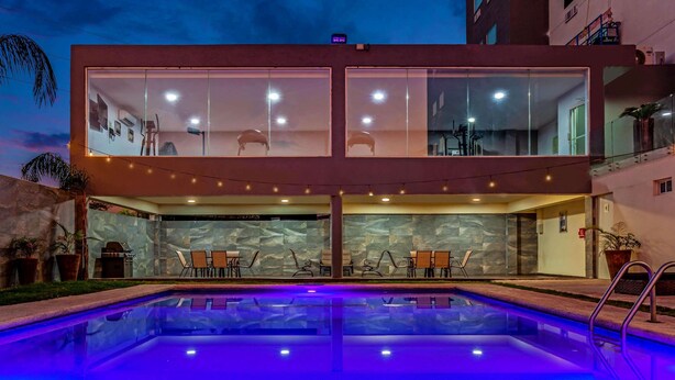 Un bâtiment moderne doté d’une façade en verre, d’une piscine éclairée de lumières bleues et d’un espace extérieur aménagé pour s’asseoir.