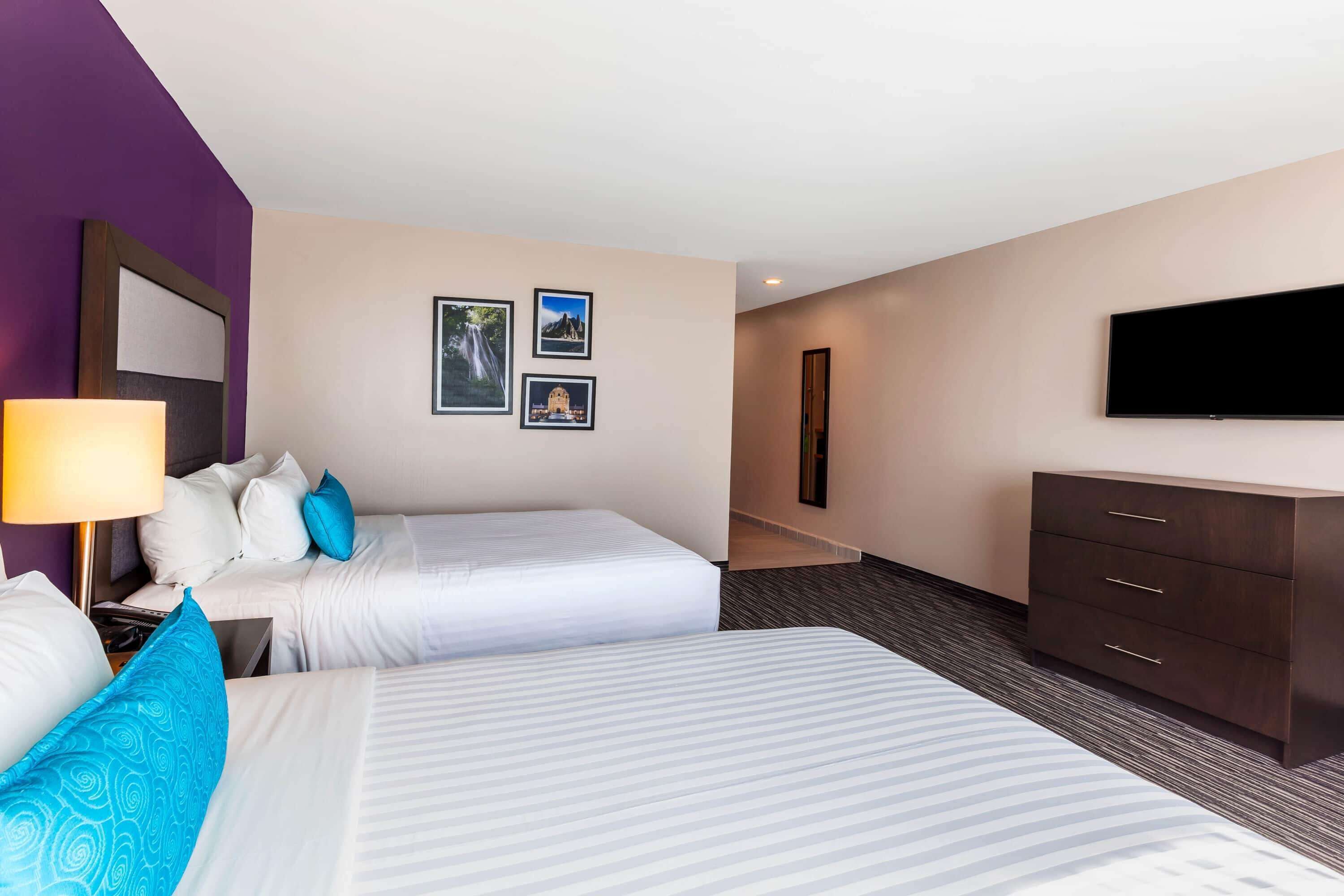 A hotel room with two beds, a TV, a dresser, and framed pictures on the wall.