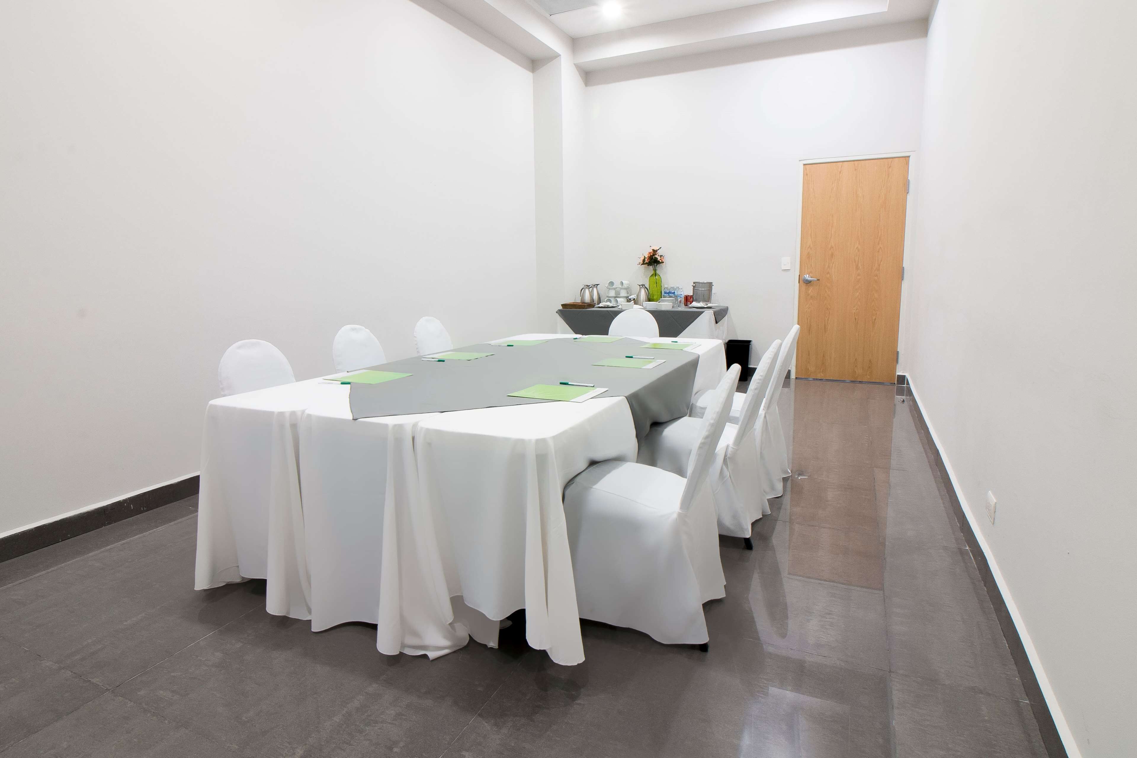 Une salle de conférence avec une table rectangulaire, des chaises blanches, une petite table sur laquelle se trouve un vase, et une porte à l’arrière-plan.
