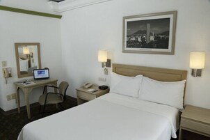 A hotel room with a bed, a desk with a laptop, a chair, a nightstand, and a wall-mounted lamp.