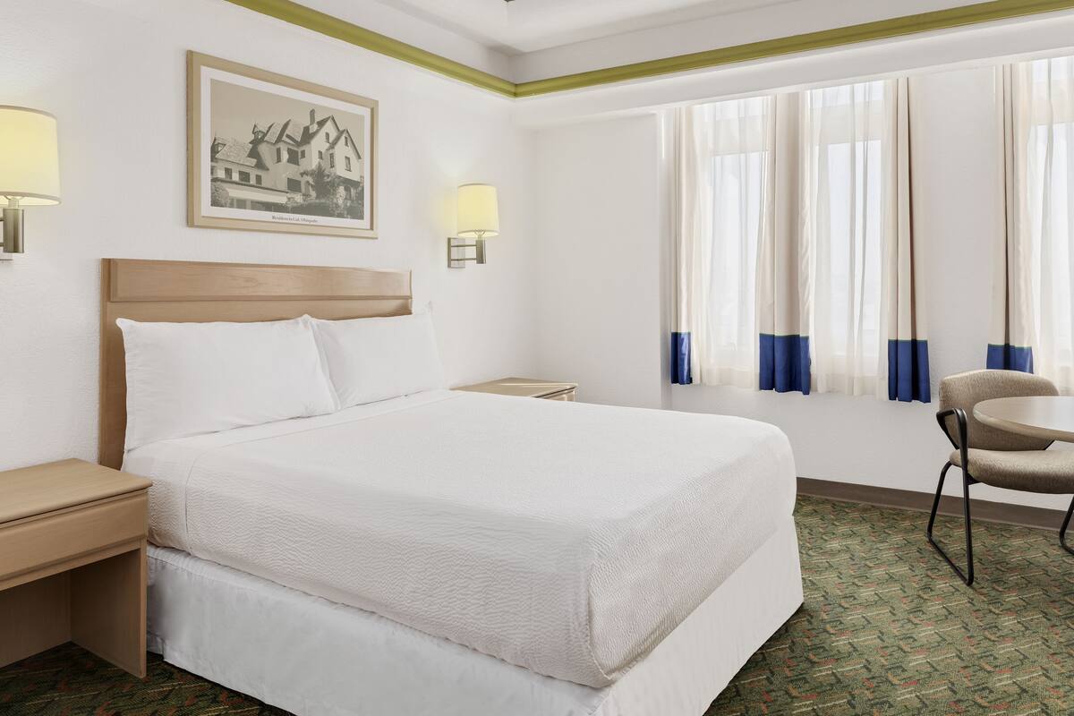 A hotel room with a large bed, two bedside tables, a chair, a small table, and a window with curtains.