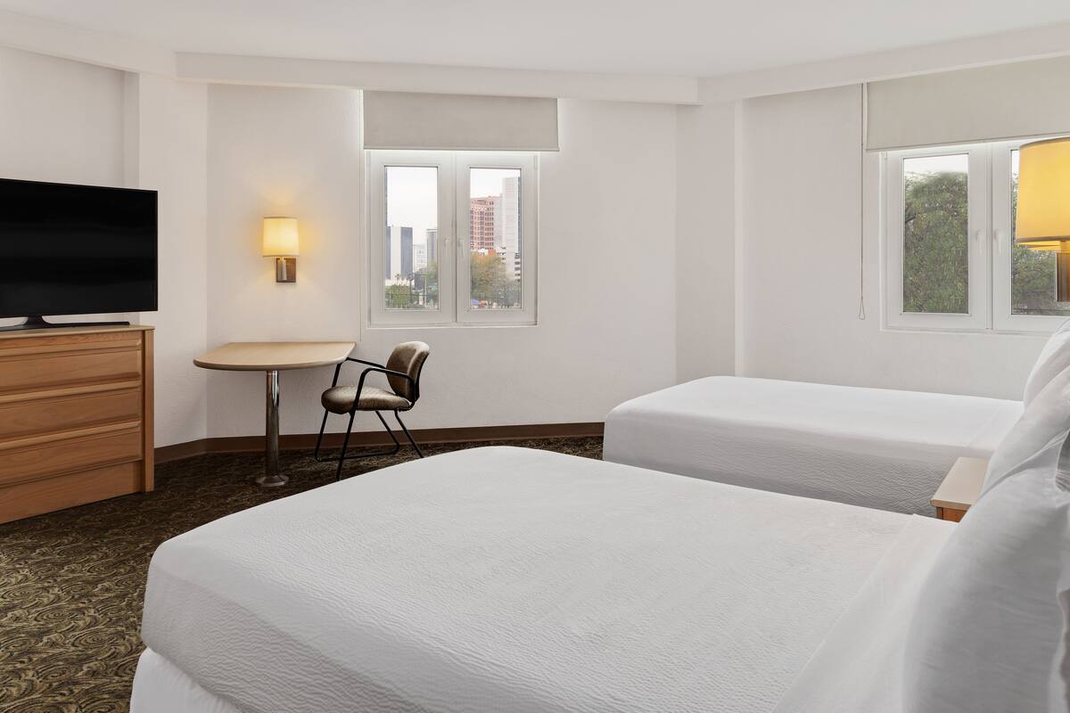 A hotel room with two beds, a TV, a small table, a chair, and a window with a view of buildings.