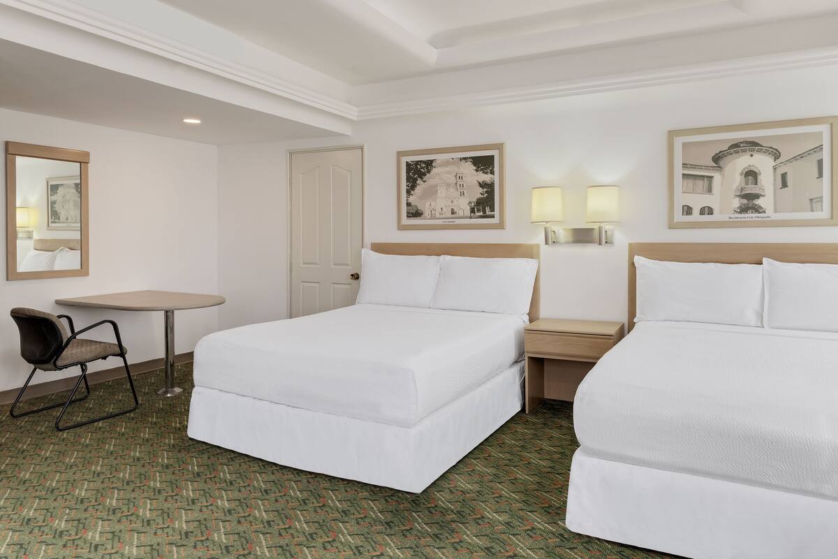 A hotel room with two beds, a small table, a chair, and two wall-mounted lamps.