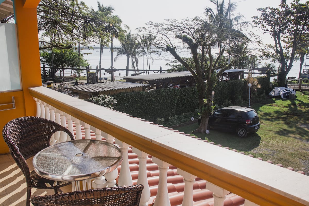 Een balkon met rieten stoelen en een tafel, met uitzicht op een strand met palmbomen en een geparkeerde auto.