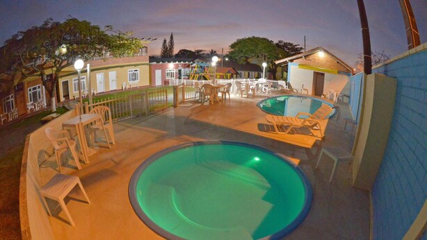 A swimming pool area with lounge chairs and tables, surrounded by a fence and buildings. - Pousada Marinamarela (Penha)